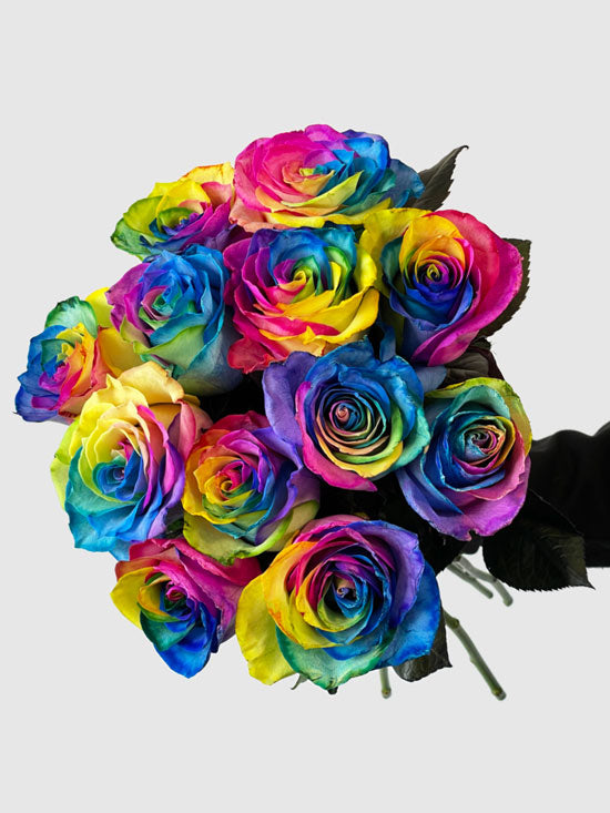 The TF Long Stem Rainbow Roses Wrapped features vibrant blue, yellow, pink, and green petals accented with lush greenery, arranged against a plain light gray background.