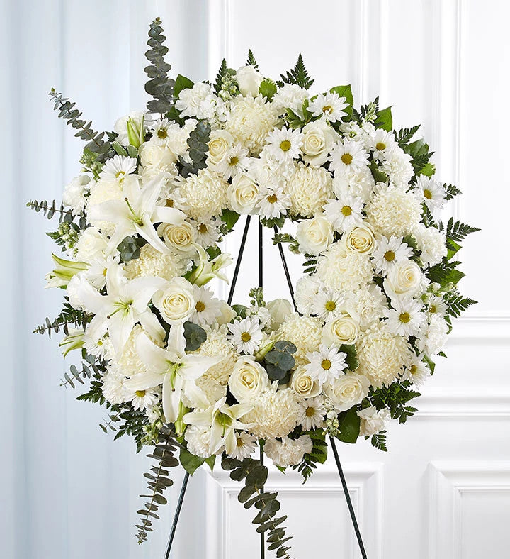 La couronne funéraire « All White » est une grande composition ronde composée de lys, de roses, de chrysanthèmes et de marguerites, agrémentée de feuillage vert et d'eucalyptus, le tout disposé sur un support métallique noir. Elle est idéale pour être exposée lors des funérailles.
