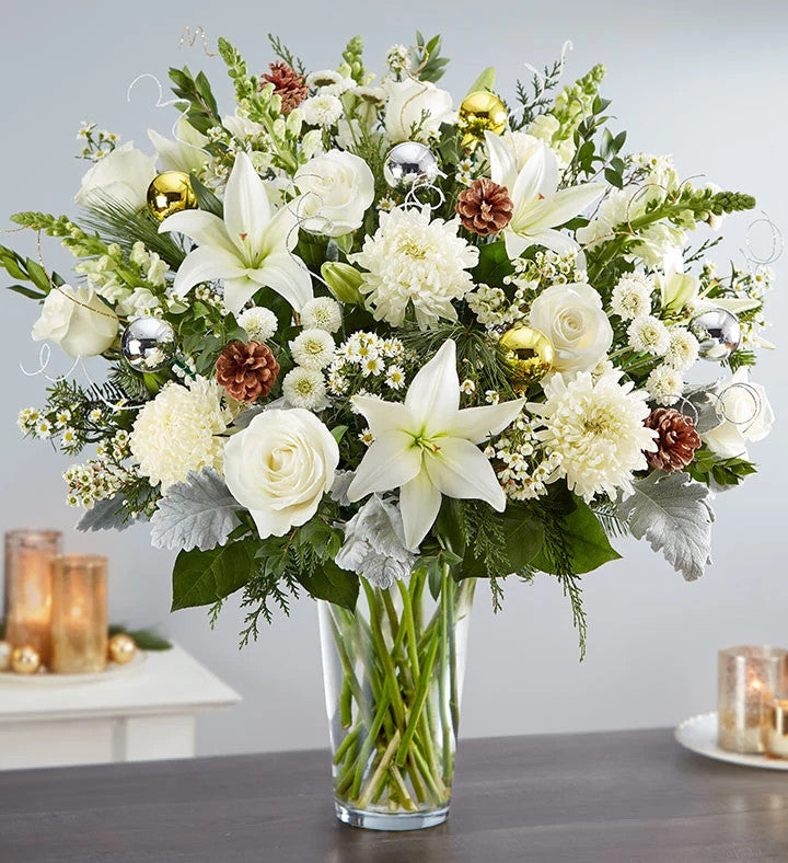 The Dazzling Winter Wonderland Flower Arrangement features white lilies, roses, chrysanthemums, pinecones, holiday greenery, and silver and gold ornaments in a glass cylinder vase—perfect for a festive table with candlelight.