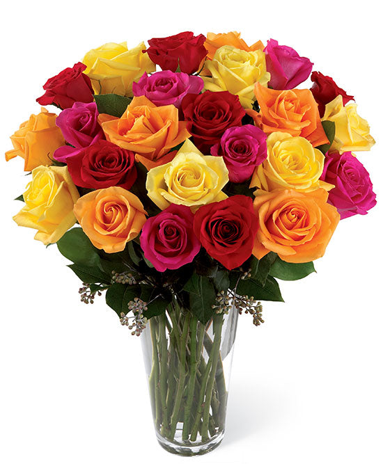 The TF Bright Spark Roses bouquet features colorful yellow, red, orange, and pink roses with lush green leaves in a clear glass vase, beautifully displayed against a simple white background.