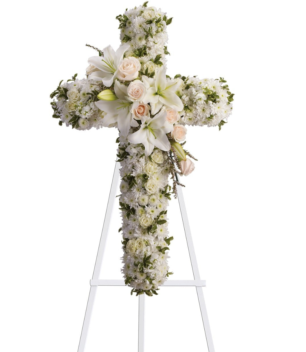 The TF "Divine Light" is a sympathy floral cross of white flowers, featuring a central bouquet of white lilies and pale pink roses, presented as an easel-mounted arrangement against a white background.