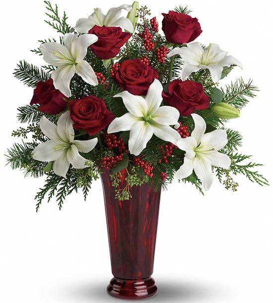 TF’s Holiday Magic is a tall red vase with a festive bouquet of red roses, white lilies, lush greenery, and red berry accents for an elegant holiday display.