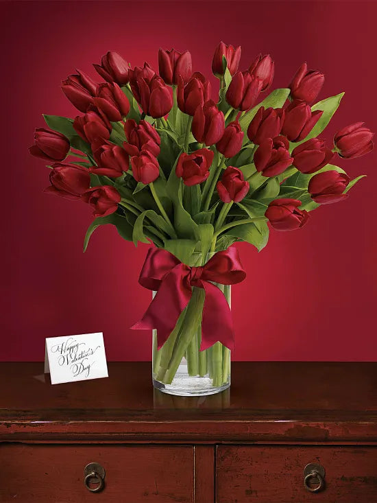 The TF Radiantly Red Tulips arrangement—vivid red tulips and lush greens tied with a red ribbon in a glass vase—is displayed on a wooden table with a Happy Mother's Day card, set against a deep red backdrop.