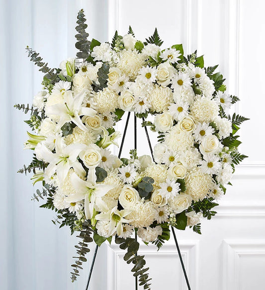 La couronne « White Solace » est une grande composition florale sur pied composée d'élégants lys blancs, de roses, de marguerites et d'une végétation luxuriante, le tout disposé sur un socle noir. Mise en valeur par un fond à panneaux blancs, cette composition funéraire entièrement blanche a été soigneusement conçue à partir de fleurs classiques afin de rendre hommage à votre proche.