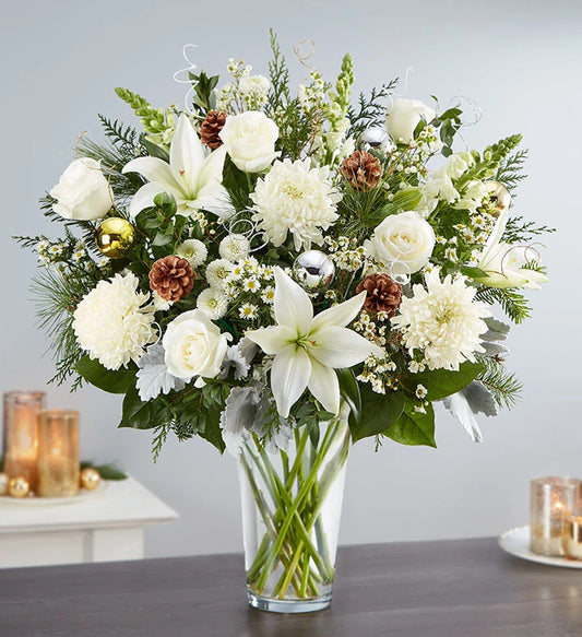 The Dazzling Winter Wonderland Flower Arrangement features white lilies, roses, chrysanthemums, pinecones, holiday greenery, and silver and gold ornaments in a glass cylinder vase—perfect for a festive table with candlelight.