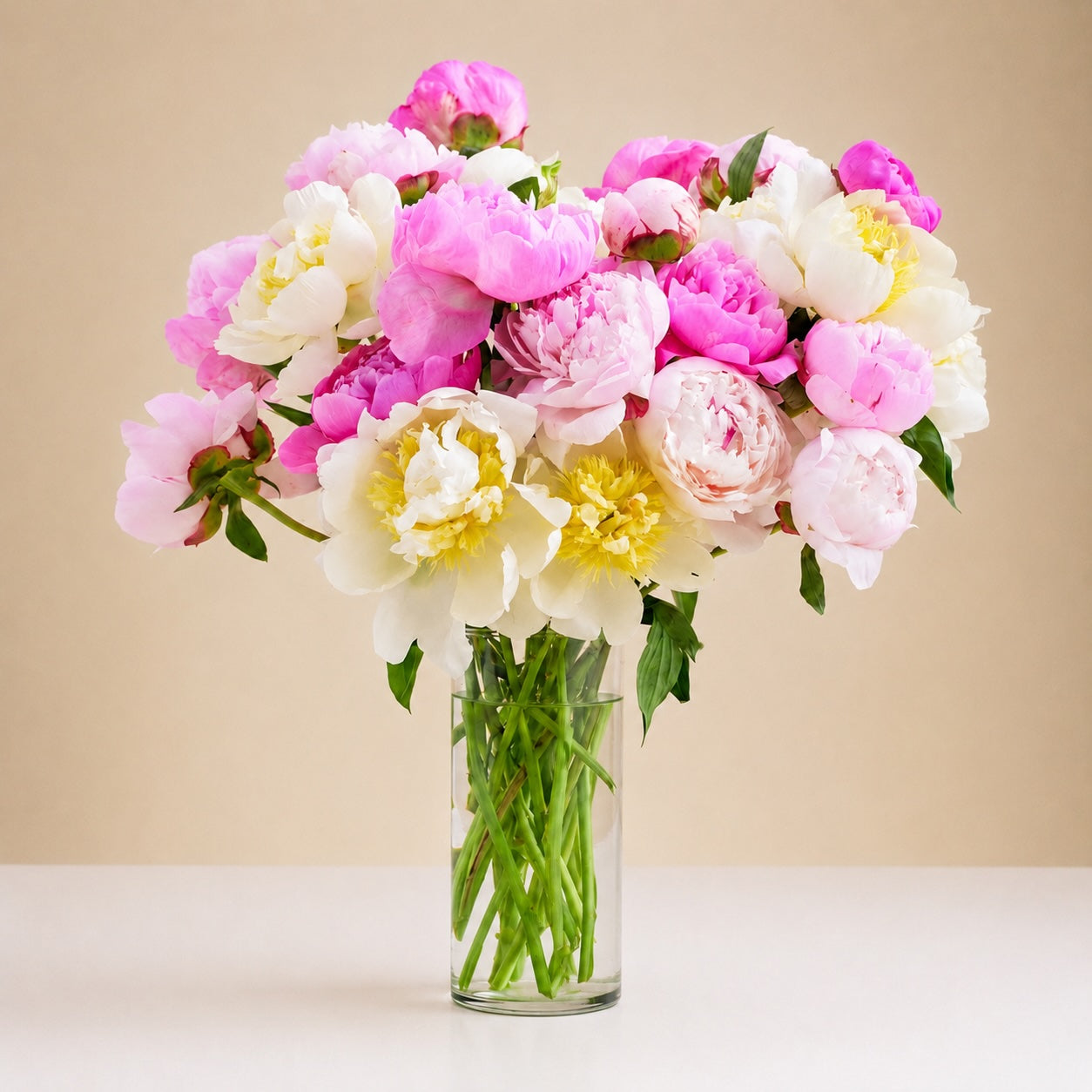 The Garden Peony Bouquet features pink and white peonies with green stems and leaves in a clear glass vase, set on a white surface against a neutral beige background.
