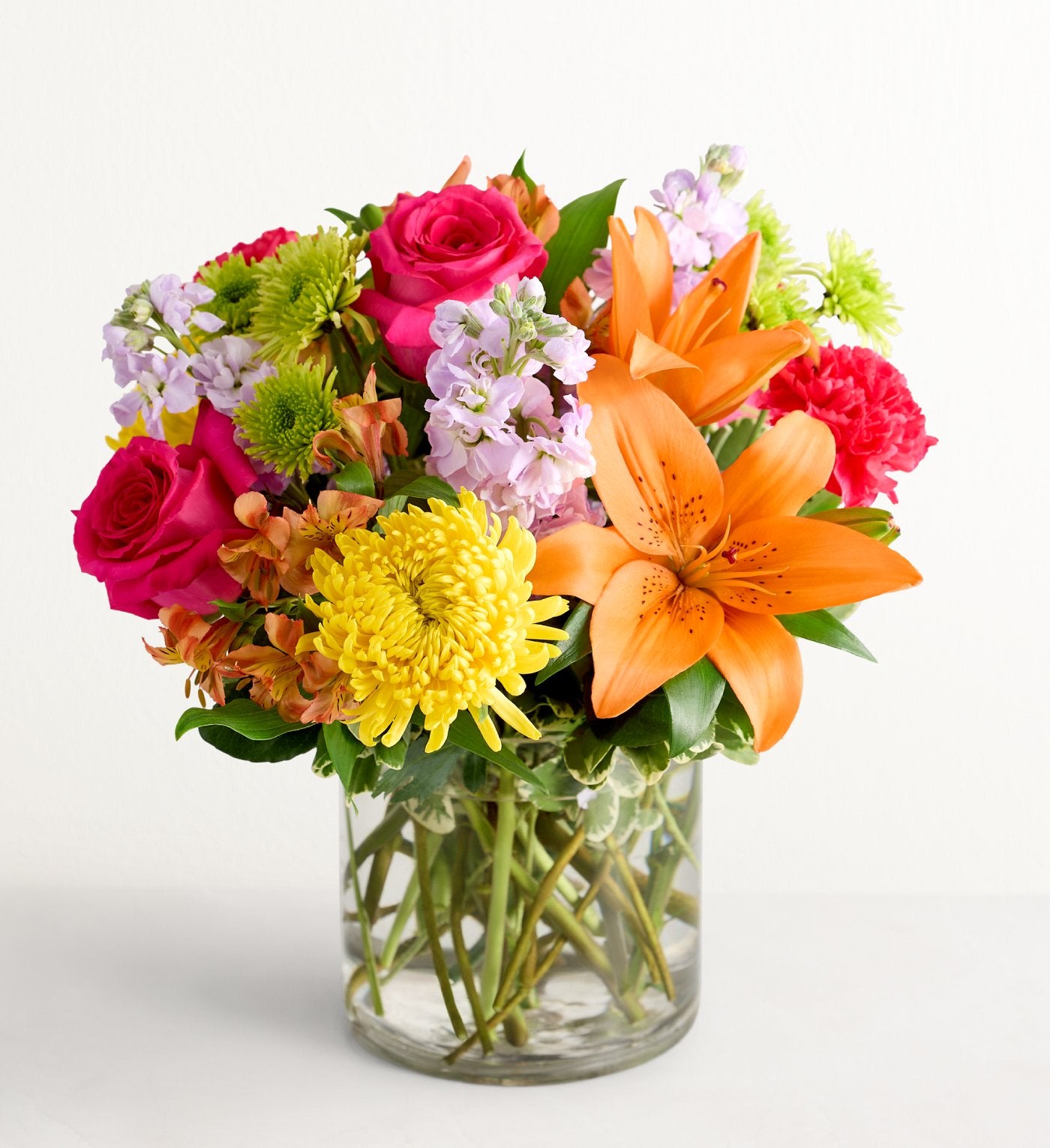 « Floral Embrace » se compose d'un vase cylindrique en verre transparent rempli de roses rose vif, de lys asiatiques orange, de chrysanthèmes jaunes, de touches de verdure et de fleurs violet clair, le tout disposé sur un fond blanc uni.