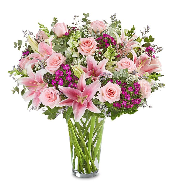 The Amaze Her Bouquet by Fleuriste Terrafolia Flowers features a glass vase filled with light pink roses, oriental lilies, deep purple flowers, white fillers, and lush greenery, beautifully arranged.