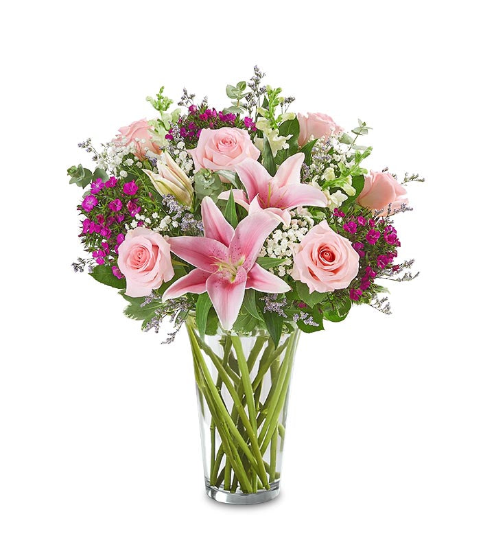 The Amaze Her Bouquet by Fleuriste Terrafolia Flowers features a glass vase filled with light pink roses, oriental lilies, deep purple flowers, white fillers, and lush greenery, beautifully arranged.