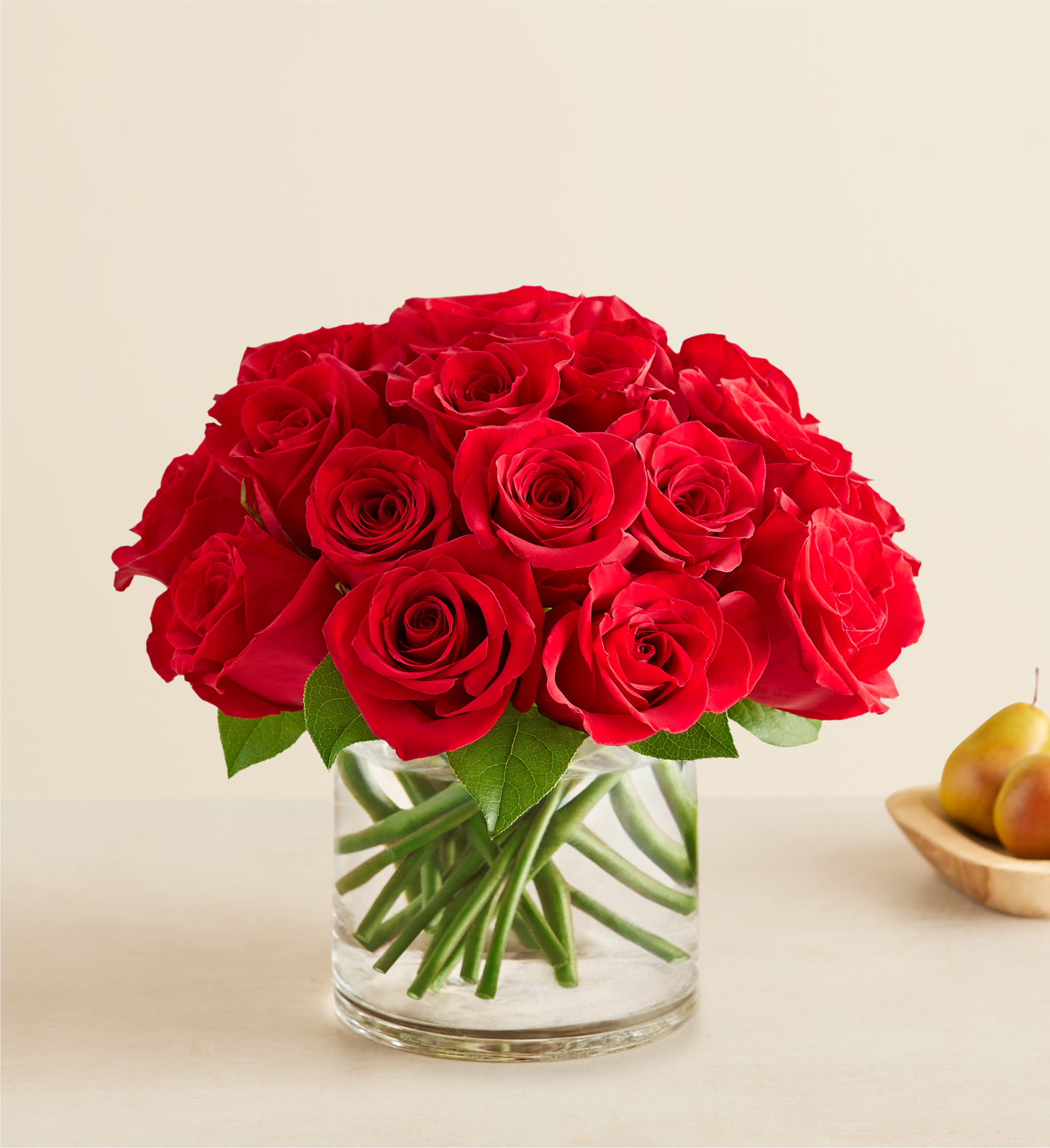 The Contemporary Red Roses features vibrant red roses with lush green leaves in a clear glass vase, set on a light surface. In the background, a wooden bowl with two pears adds charm to the elegant arrangement.