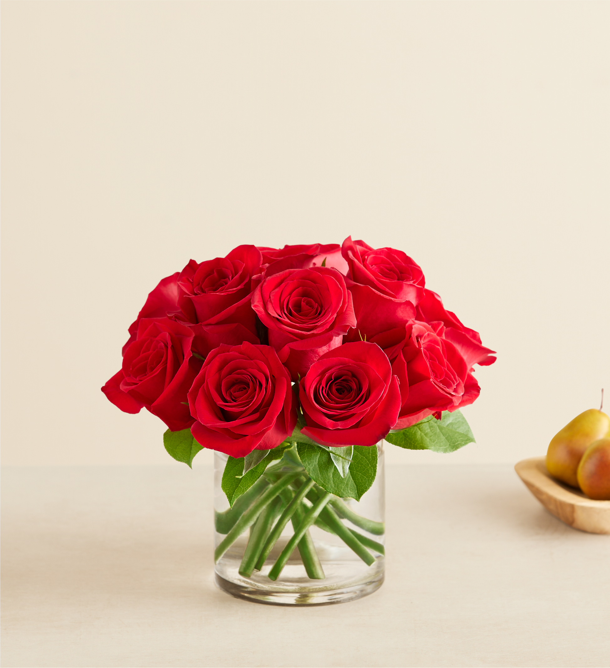 The Contemporary Red Roses features vibrant red roses with lush green leaves in a clear glass vase, set on a light surface. In the background, a wooden bowl with two pears adds charm to the elegant arrangement.