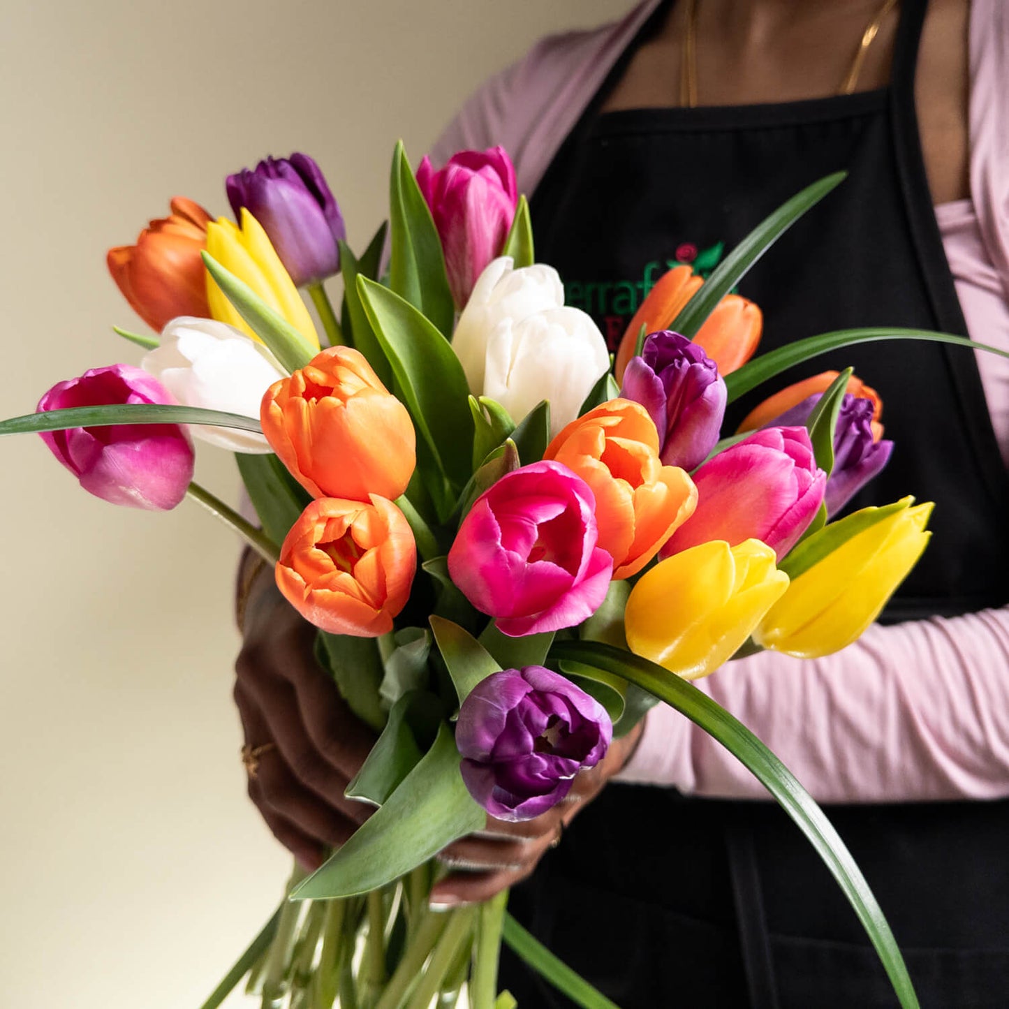 The TF Colorful Mixed Tulips Wrapped bouquet, featuring vibrant orange, yellow, purple, and white tulips, rests on a white surface with a simple beige background.