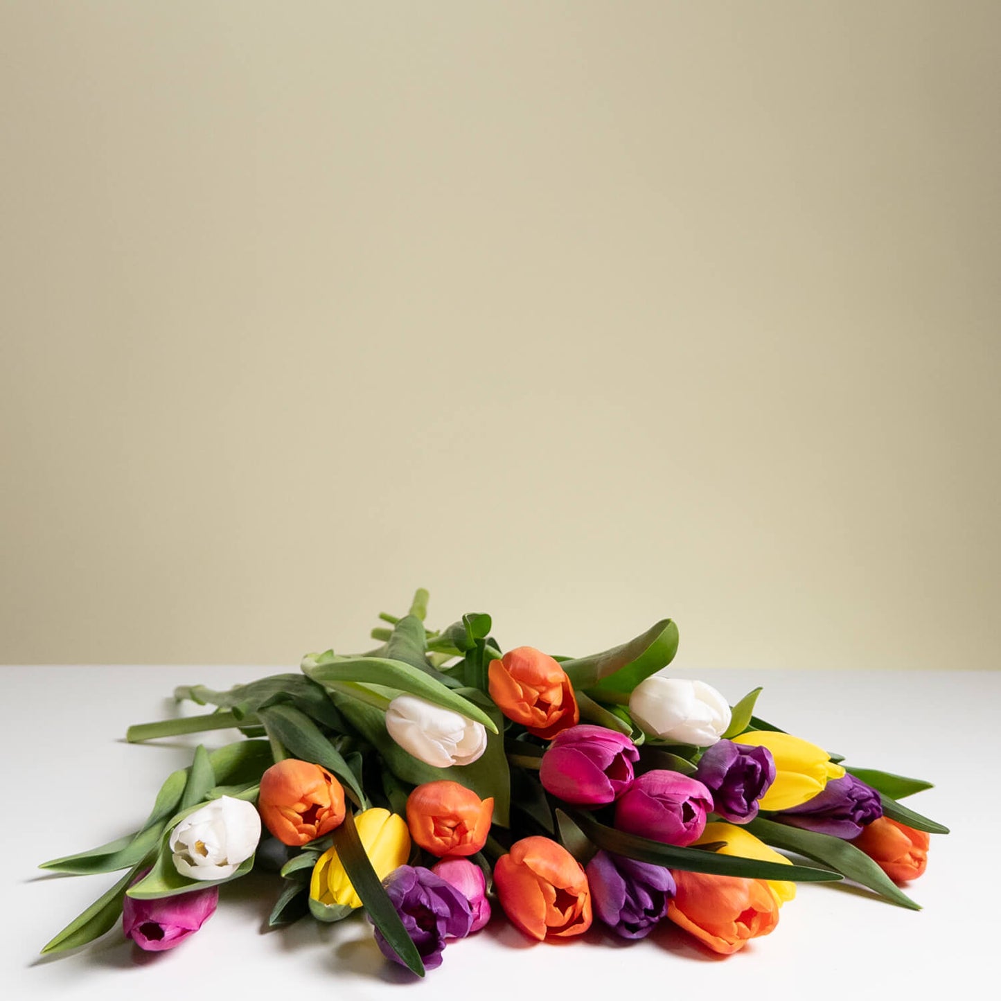 The TF Colorful Mixed Tulips Wrapped bouquet, featuring vibrant orange, yellow, purple, and white tulips, rests on a white surface with a simple beige background.