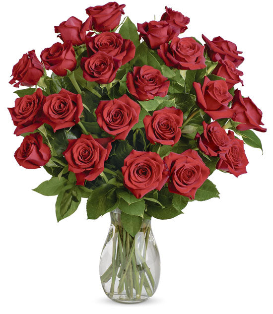 A bouquet of TF Long Stem Red Roses with green leaves is arranged in a clear vase on a light table. Two red candles—one lit, one unlit—in the background bring added warmth beside the roses.