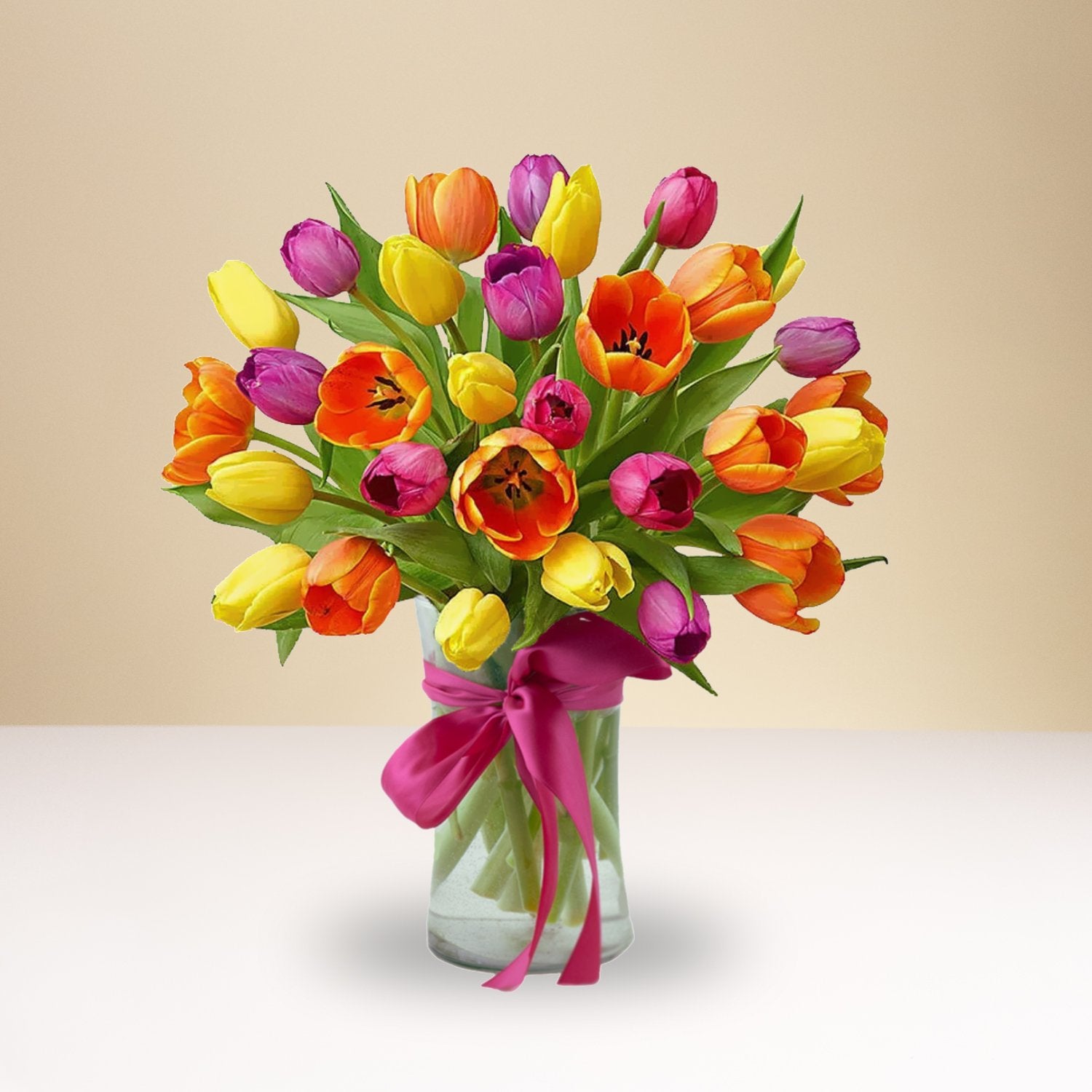 The Colorful Mixed Tulips bouquet features yellow, orange, pink, and purple tulips artfully arranged in a glass vase with a bright pink ribbon, set against a soft beige background.