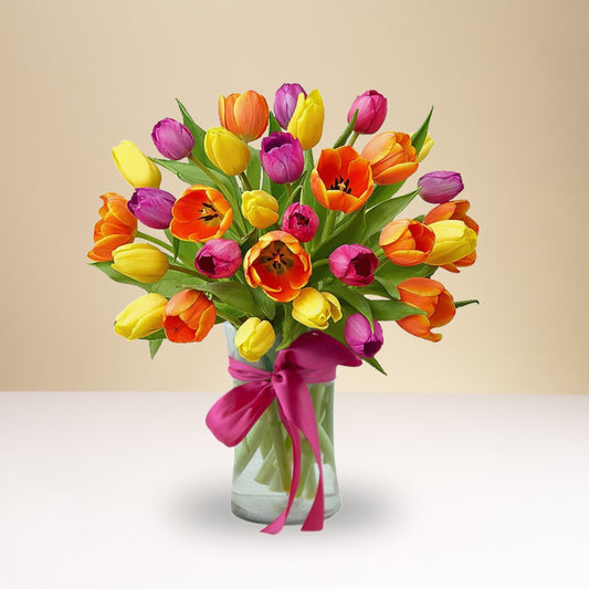 The Colorful Mixed Tulips bouquet features yellow, orange, pink, and purple tulips artfully arranged in a glass vase with a bright pink ribbon, set against a soft beige background.