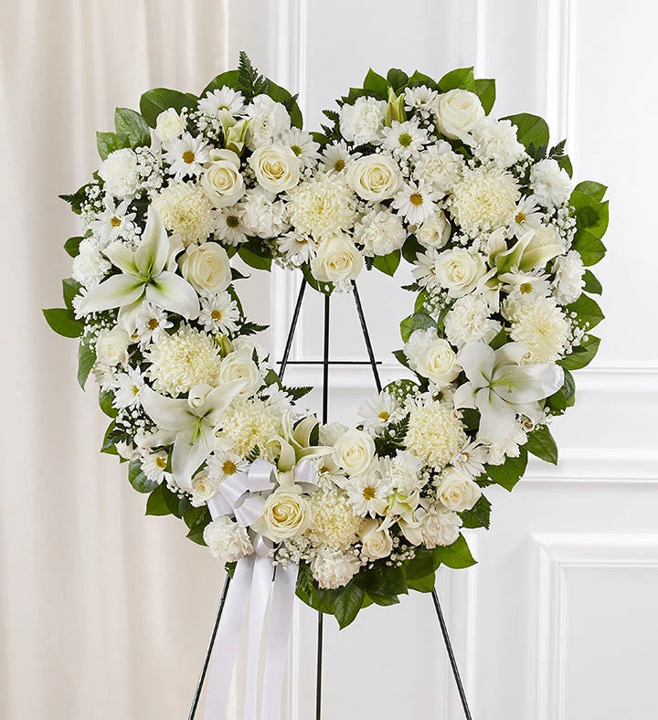 La composition « Always Remember » se compose d'un grand cœur de roses blanches, de lys, de marguerites et de chrysanthèmes, agrémenté de feuillage vert, disposé sur un socle noir devant un mur blanc, et orné d'un ruban blanc à sa base.