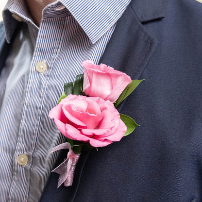 The Velvet Honor Boutonniere features two pink lisianthus flowers with green leaves, tied with a light pink ribbon against a white background—an elegant choice for formal events.