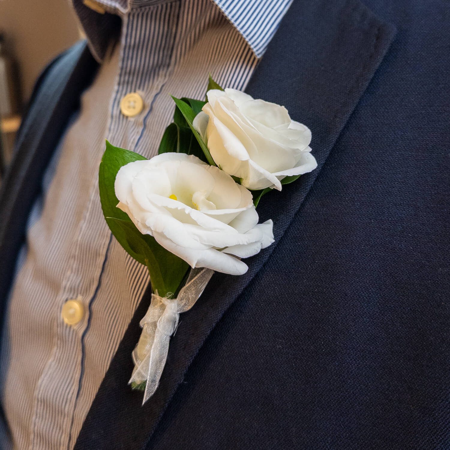 The Noble Stem Boutonniere features two white lisianthus blooms with yellow centers and green leaves, elegantly arranged—an ideal accessory for formal events—shown against a plain white background.
