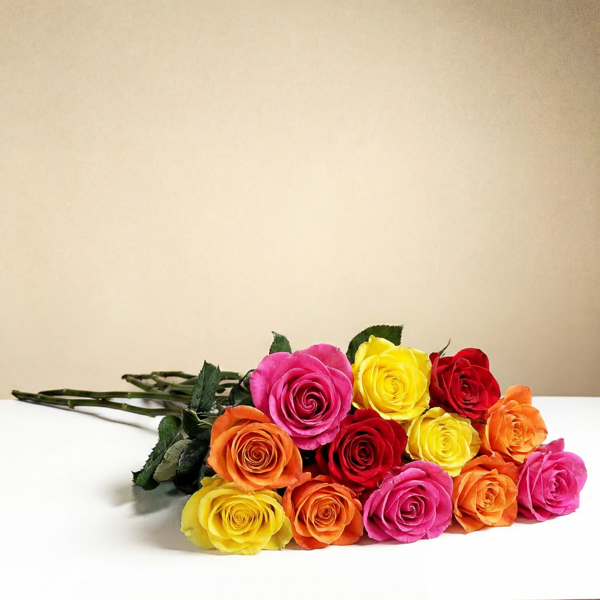The TF Bright Spark Roses Wrapped features a romantic bouquet of vibrant pink, yellow, orange, and red roses arranged on a white surface with a plain beige background.