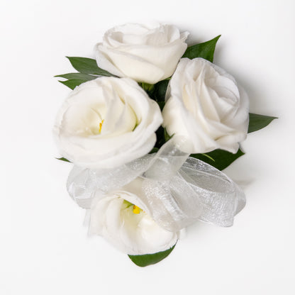 The White Muse Wristlet features three hand-crafted white roses with green leaves, tied with a sheer white ribbon—an elegant accessory perfect for weddings or formal occasions. Displayed on a white background.
