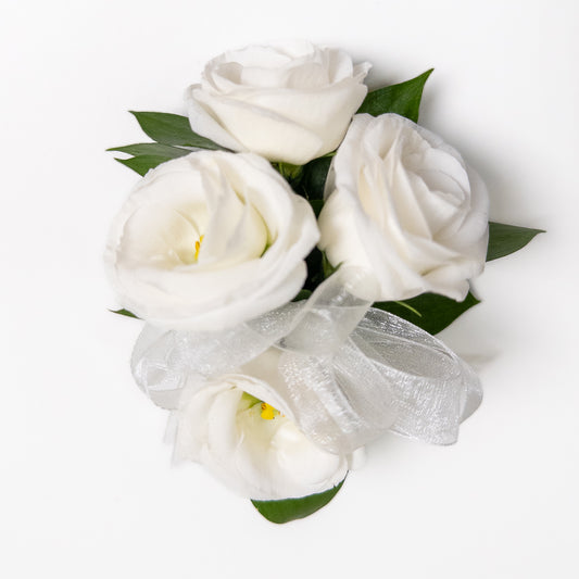The White Muse Wristlet features three hand-crafted white roses with green leaves, tied with a sheer white ribbon—an elegant accessory perfect for weddings or formal occasions. Displayed on a white background.