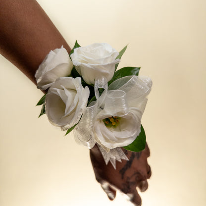 The White Muse Wristlet features three hand-crafted white roses with green leaves, tied with a sheer white ribbon—an elegant accessory perfect for weddings or formal occasions. Displayed on a white background.