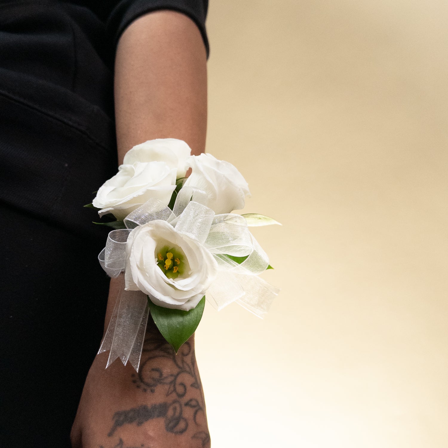 The White Muse Wristlet features three hand-crafted white roses with green leaves, tied with a sheer white ribbon—an elegant accessory perfect for weddings or formal occasions. Displayed on a white background.