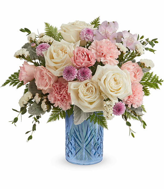 The Blue Belle Bouquet features white roses, pink carnations, purple button poms, and greenery, artfully arranged in our signature Blue Belle Vase.