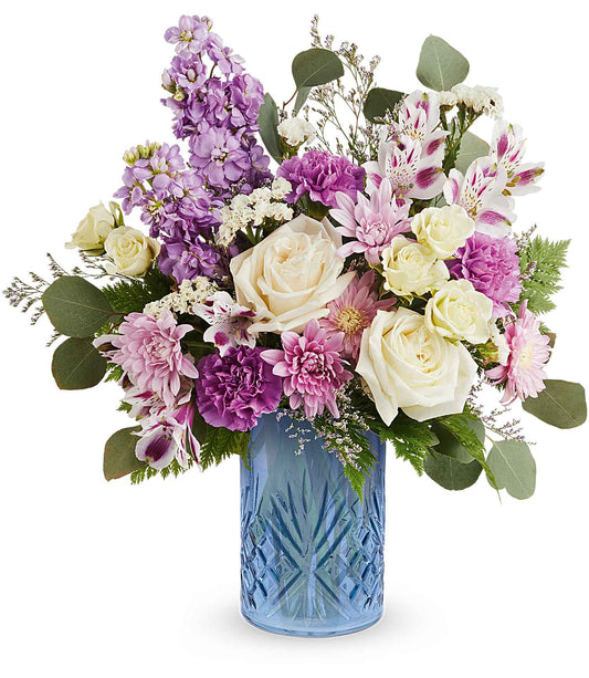 The Blue Bliss Bouquet features white roses, pink carnations, lavender, greenery, and small white blossoms stylishly arranged in a striking Blue Belle Vase.
