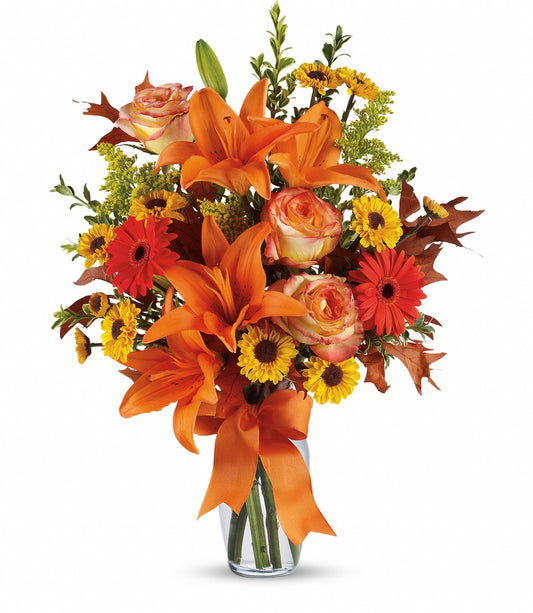 The Burst of Autumn by TF features a clear glass vase filled with Asiatic lilies, red roses, yellow daisies, autumn leaves, and an orange ribbon for a vibrant fresh flower display.