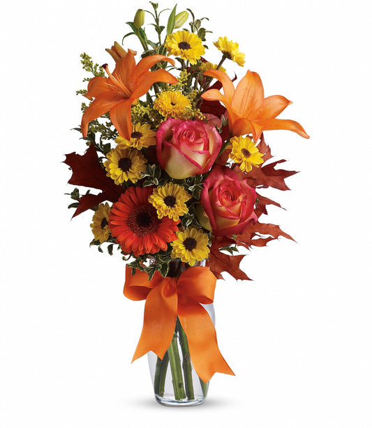 The Burst of Autumn by TF features a clear glass vase filled with Asiatic lilies, red roses, yellow daisies, autumn leaves, and an orange ribbon for a vibrant fresh flower display.