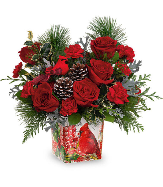 The Cardinal Cheer Bouquet by Fleuriste Terrafolia Flowers is a festive arrangement with red roses, red carnations, pine branches, pinecones, and greenery in a square vase decorated with a red cardinal and holiday designs.