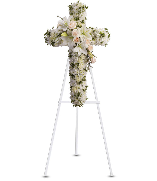 The TF "Divine Light" is a sympathy floral cross of white flowers, featuring a central bouquet of white lilies and pale pink roses, presented as an easel-mounted arrangement against a white background.
