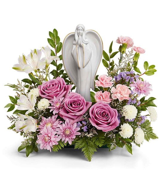 The Ever So Loved Bouquet features lavender roses, pink carnations, white lilies, small white flowers, purple accents, and green foliage arranged around a white Angel of Grace keepsake figurine.