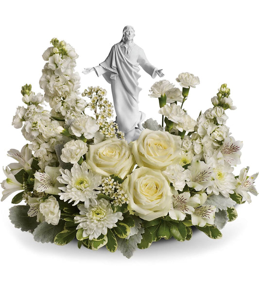 The TF Forever Faithful Bouquet showcases pure white roses, chrysanthemums, carnations, lush greenery, and a Sacred Grace keepsake statue of Jesus with outstretched arms at the center.