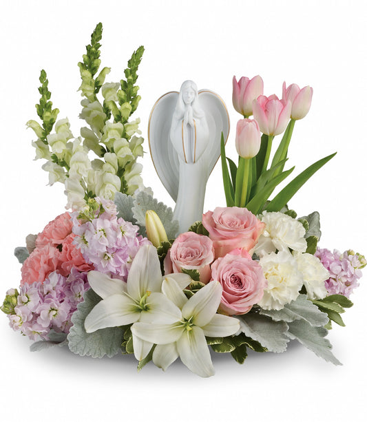 The Garden Of Hope Bouquet features white lilies, pink roses, light pink flowers, carnations, and tulips surrounding a porcelain angel figurine with pastel tones and green and silver foliage.