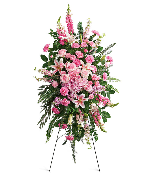 The Glorious Farewell Spray is a pink floral easel arrangement with roses, lilies, carnations, snapdragons, and greenery on a sleek metal stand against a white background.