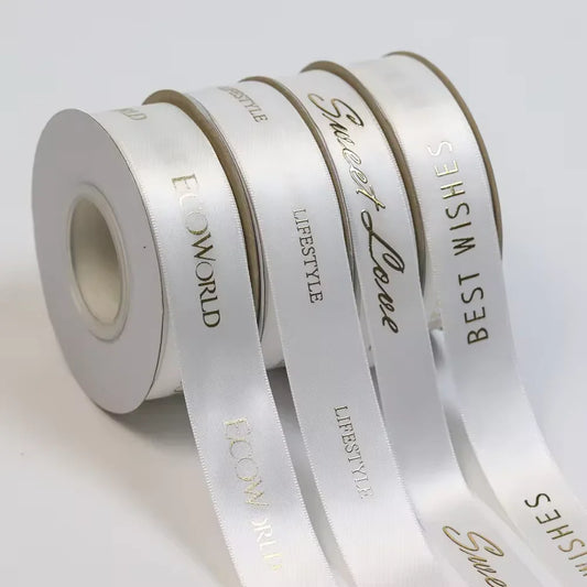 Quatre bobines de ruban pour bannières imprimées « Add », en satin blanc avec des inscriptions dorées — « ECOWORLD », « LIFESTYLE », « Sweet Love » et « BEST WISHES » — sont disposées en rangée.