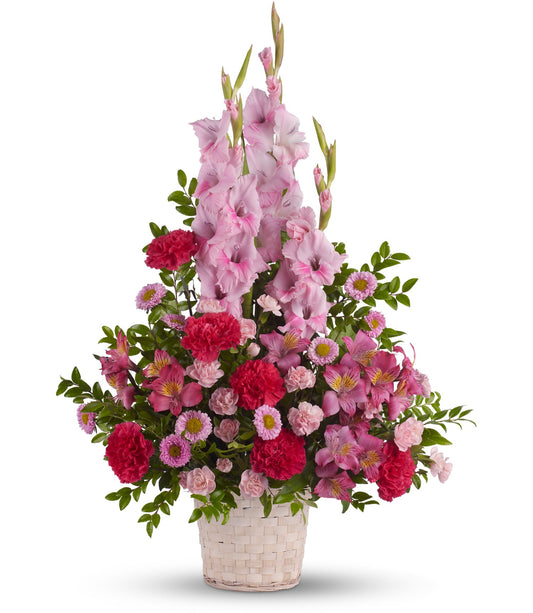 The TF "Heavenly Heights" is a white woven basket filled with pink gladiolus, carnations, alstroemeria, purple asters, and lush green foliage.