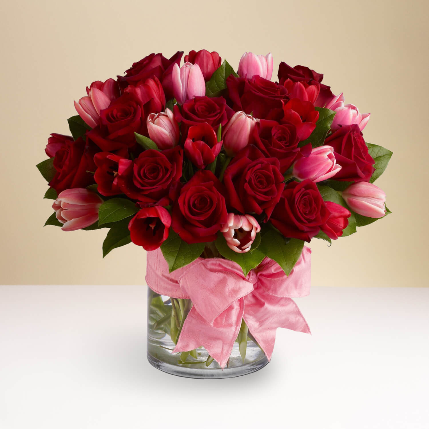 The TF "P.S. I Love You" features a clear glass vase of red roses, pink and red tulips, lush green leaves, and a big pink ribbon bow, arranged on a white surface with a beige background for an elegant display.