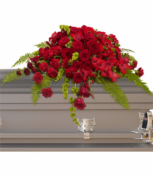 A gray closed casket with silver handles features the Red Rose Sanctuary Casket Spray, a saddle-mounted arrangement of red roses, gladioli, and green ferns on top.