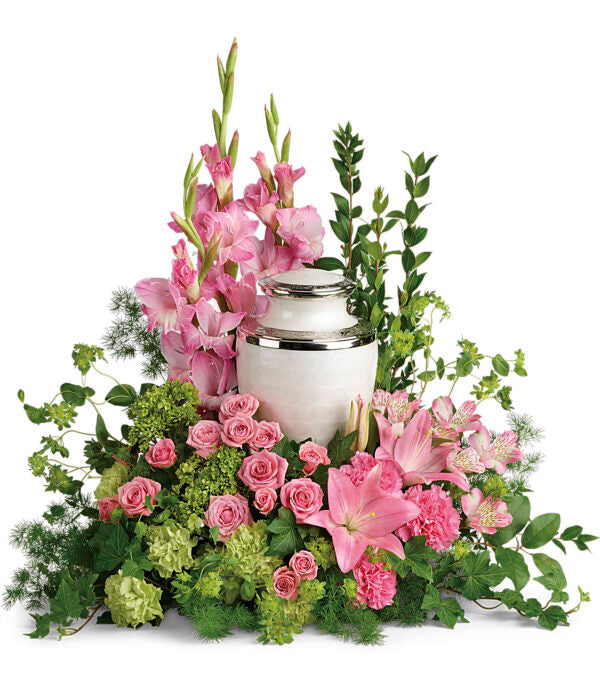 The Sacred Solace Cremation Tribute by Fleuriste Terrafolia Flowers features a white urn with silver trim, surrounded by pink lilies, roses, gladiolus, carnations, spray roses, green hydrangea, and lush greenery.