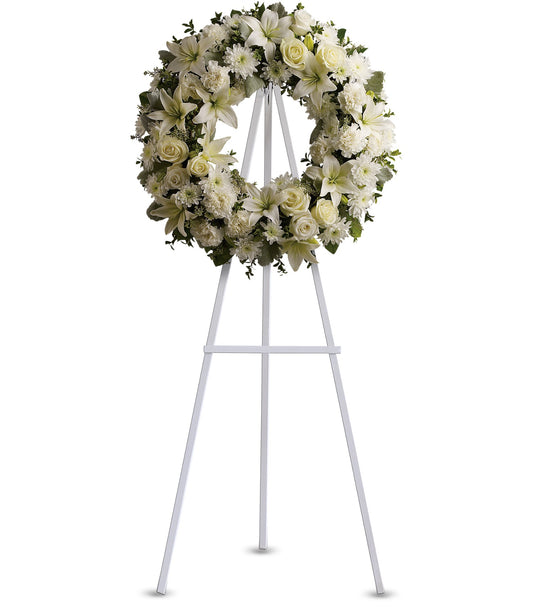 The TF Serenity Wreath, featuring fresh white roses and lilies, is displayed on a tall white metal easel against a plain white background.