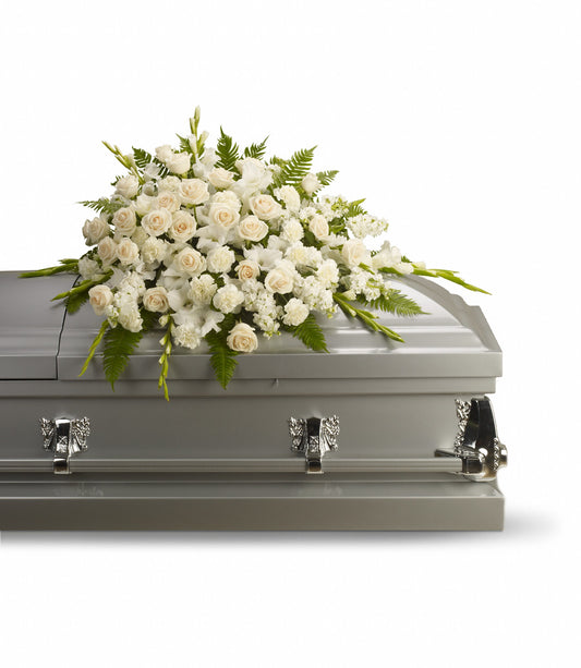 The Silken Serenity Casket Spray features a lush floral arrangement of white roses, gladioli, and greenery atop a closed silver casket, all set against a plain white background.