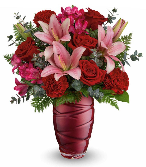 The TF Swirling Embrace Bouquet features pink asiatic lilies, red roses, red carnations, and lush greenery in a vibrant red vase for a stunning floral arrangement.