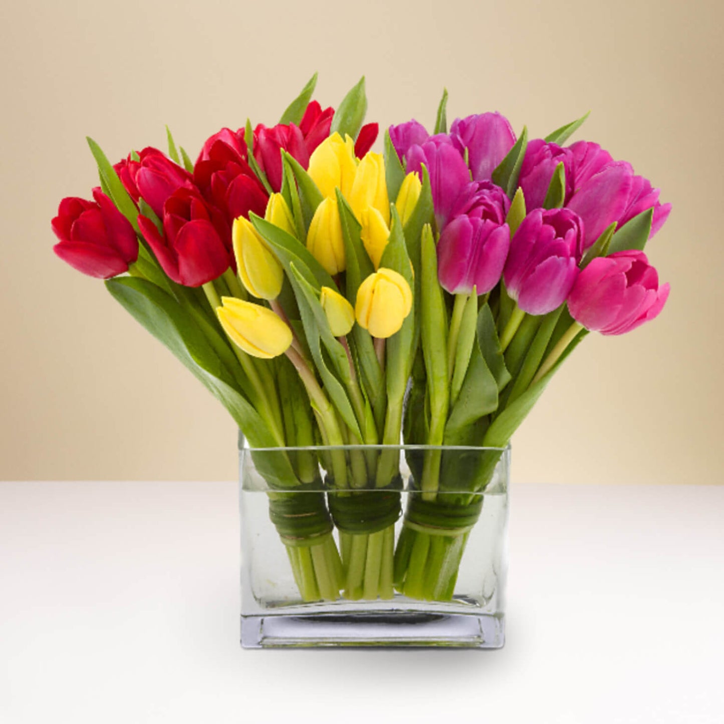 The Tulips Together arrangement features a clear rectangular glass vase filled with vibrant red, yellow, and purple tulips with green stems and leaves, displayed against a neutral beige background.